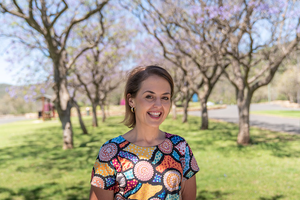 A person in a colourful top stands in a park, smiling at the camera with a row of trees behind them.