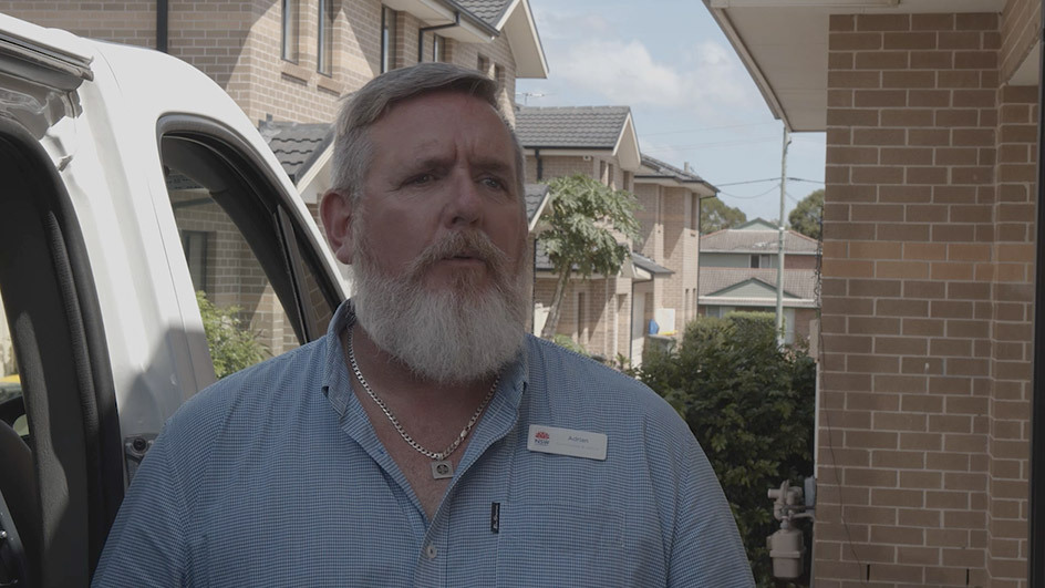 A person with a beard talking to the camera, with a row of houses behind them.