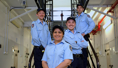 Four people in blue Correctional Officer uniforms stand together on a metal staircase, facing the camera.