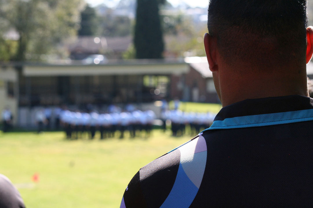 The back of the head of a person who is looking towards a group of people in blue Correctional Officer uniforms, in a grassy open space, out of focus in the distance.