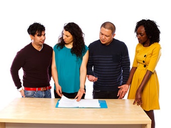 A group of people standing at a table, reading a document together. 