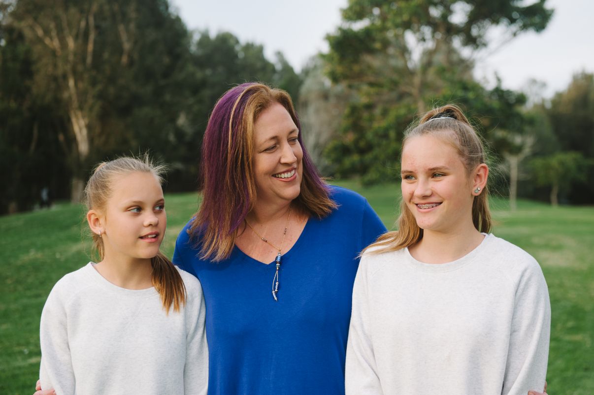 A female adult with coloured hair smiling whilst standing with two children who also look happy