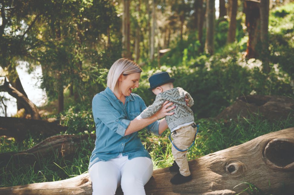 A female adult sitting on a log holding up a male toddler smiling and playing
