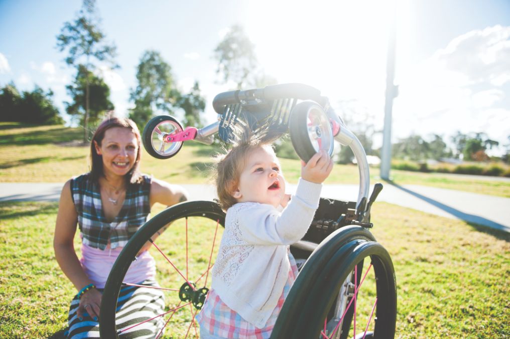 An adult female watching a female toddler playing with a bicycle both appear to be happy smiling