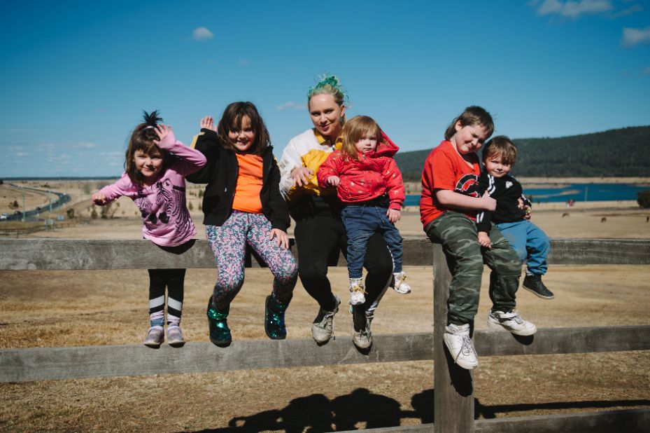 A female with green hair sitting on a fence with five young children all smiling and some waving