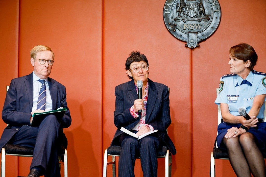 Government Panel speaking on the stage while seated. The person in the middle is speaking into the microphone while the other two people are listening.