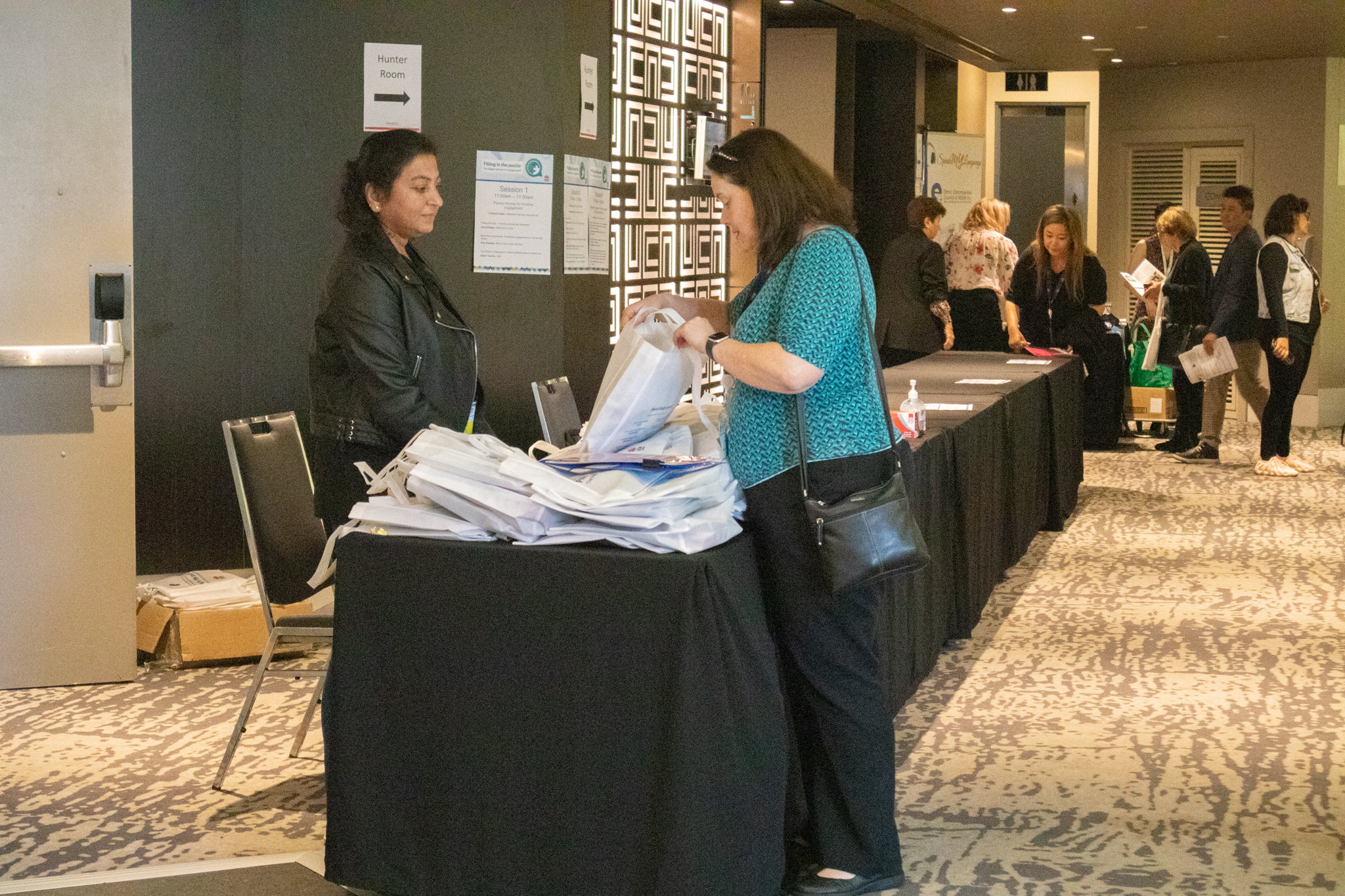 A person standing at a table, looking inside a conference bag, with another person looking on.