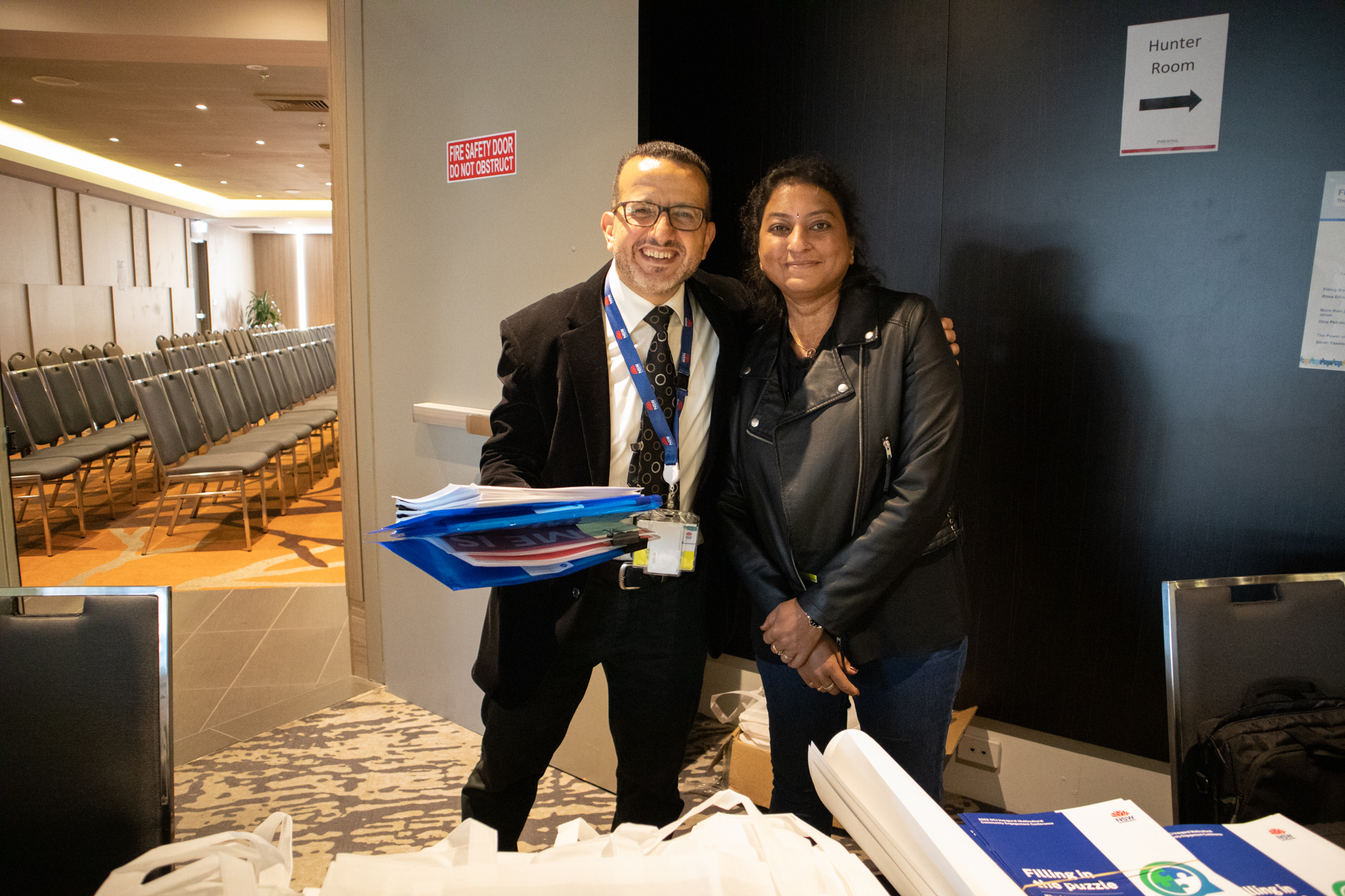 Two people standing closely together behind a conference table, holding a handful of folders, and smiling at the camera.