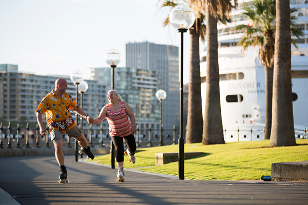 Andrew and Debbie hold hands as they roller skate together down a city path on a sunny day, with apartment buildings and palm trees behind them.