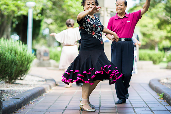 Two ballroom dancers wearing black and pink hold hands and dance together, on a paved path surrounded by trees, with other dancers in the background.