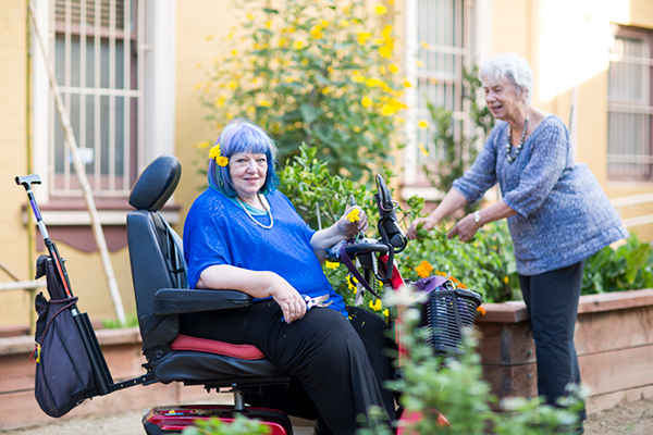 Christine sitting in a mobility power chair, holding flowers and with flowers in her hair. Another person standing beside her tends to the garden.