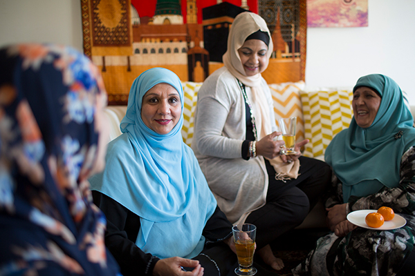 Mahboha sitting on a couch with three other women, all wearing hijabs, holding drinks and food. Mahboha looks at the camera and the other women smile.