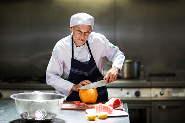 Merv Parker in a commercial kitchen, wearing chef's clothing and an apron, cutting a pumpkin.