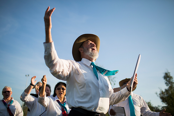 Simply Voices choir wearing white shirts and coloured ties, their arms in the air as they sing against the blue sky.