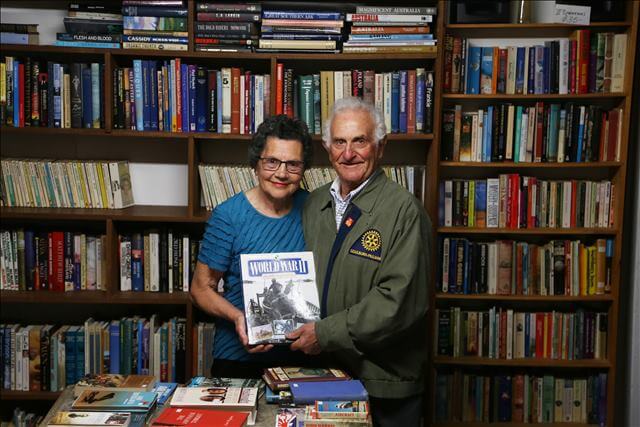 Adriana and Tony standing together in a room full of books. They stand close together, holding a World War II book. There is a wall of books behind them and a table with books on it in front of them. 