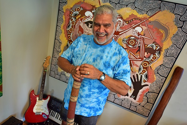 Pastor Russell Saunders OAM standing in front of a painting, guitar and keyboard, holding a didgeridoo and smiling at the camera.