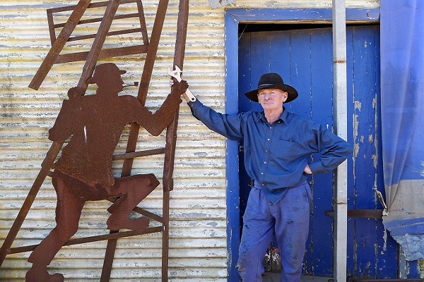 Mark Oats standing outside in front of a blue wooden door and corrugated wall, against which is a flat sculpture of a man climbing a ladder.
